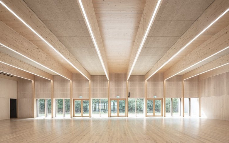 Im Innen der Kulturhalle dominieren helle Sichtholzflächen, der Blick schweift durch die fast gebäudebreite Glasfront in den direkt angrenzenden Bienwald.  Bild: AV1 Architekten GmbH, Kaiserslautern