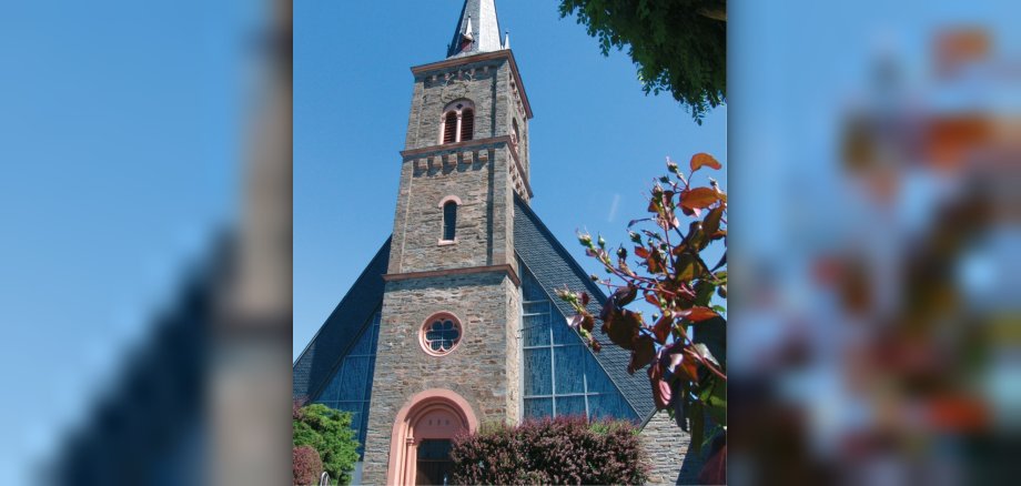 Portal und Kirchturm der katholischen Pfarrkirche St. Andreas der Ortsgemeinde Longkamp vor blauem Himmel.