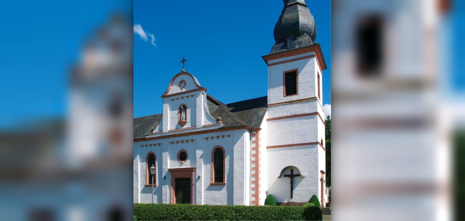  Eingang und Kirchturm der Pfarrkirche Sankt Hubertus vor blauem Himmel.